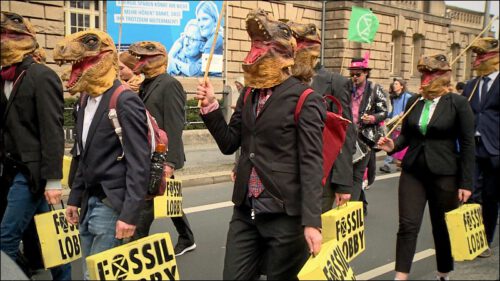 In der satirischen Demo der Superreichen: An­zug­trä­ge­r*in­nen in Dino-Gummimasken und mit gelben Aktenkoffern in den Händen – Vertreter der fossilen Lobby