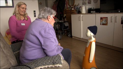 Eine Pflegerin und eine ältere Bewohnerin sitzen im Zimmer eines Seniorenheims und schauen den kleinen Sozialroboter Gottfried an – ein orangefarbener Roboter mit Strickmütze, der einmal pro Woche in der Einzelbetreuung eingesetzt wird.