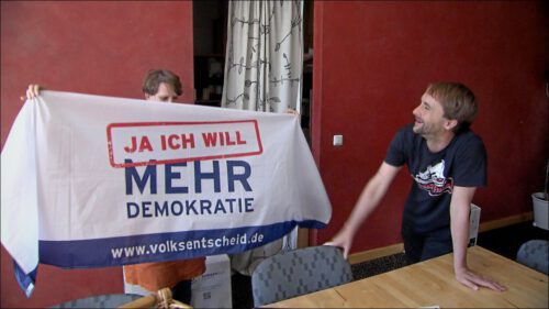 Christian König und Aline Koeppen im Berliner Büro von Mehr Demokratie e.V. halten ein Banner mit der Aufschrift "Ja ich will Mehr Demokratie – www.volksentscheid.de" hoch.