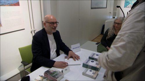 Ralf-Uwe Beck sitzt an einem Bücherverkaufstisch und signiert ein Exemplar seines Buchs "Der Souverän".