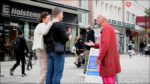 Der langjährige OMNIBUS-Fahrer Werner Küppers, in auffälligem rot-gemustertem Mantel und pinker Hose, spricht in einer Fußgängerzone mit zwei Passant*innen. Neben ihm ein Schild: »Rettet den Bürgerentscheid – Volksinitiative Bürgerbegehren in Schleswig-Holstein«.