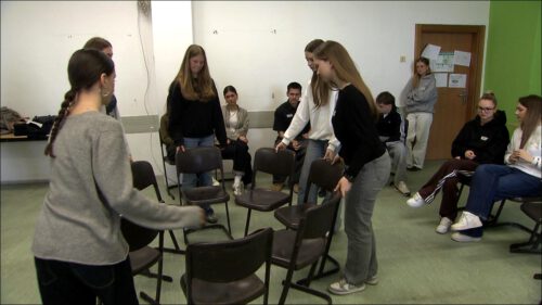 Jugendliche stellen in einem Klassenzimmer des Fürther Helene-Lange-Gymnasiums Stühle zu einem Kreis auf, im Hintergrund sitzen weitere Schüler:innen und beobachten die Übung – Szene vom Schulprojekt "wo-hin?".
